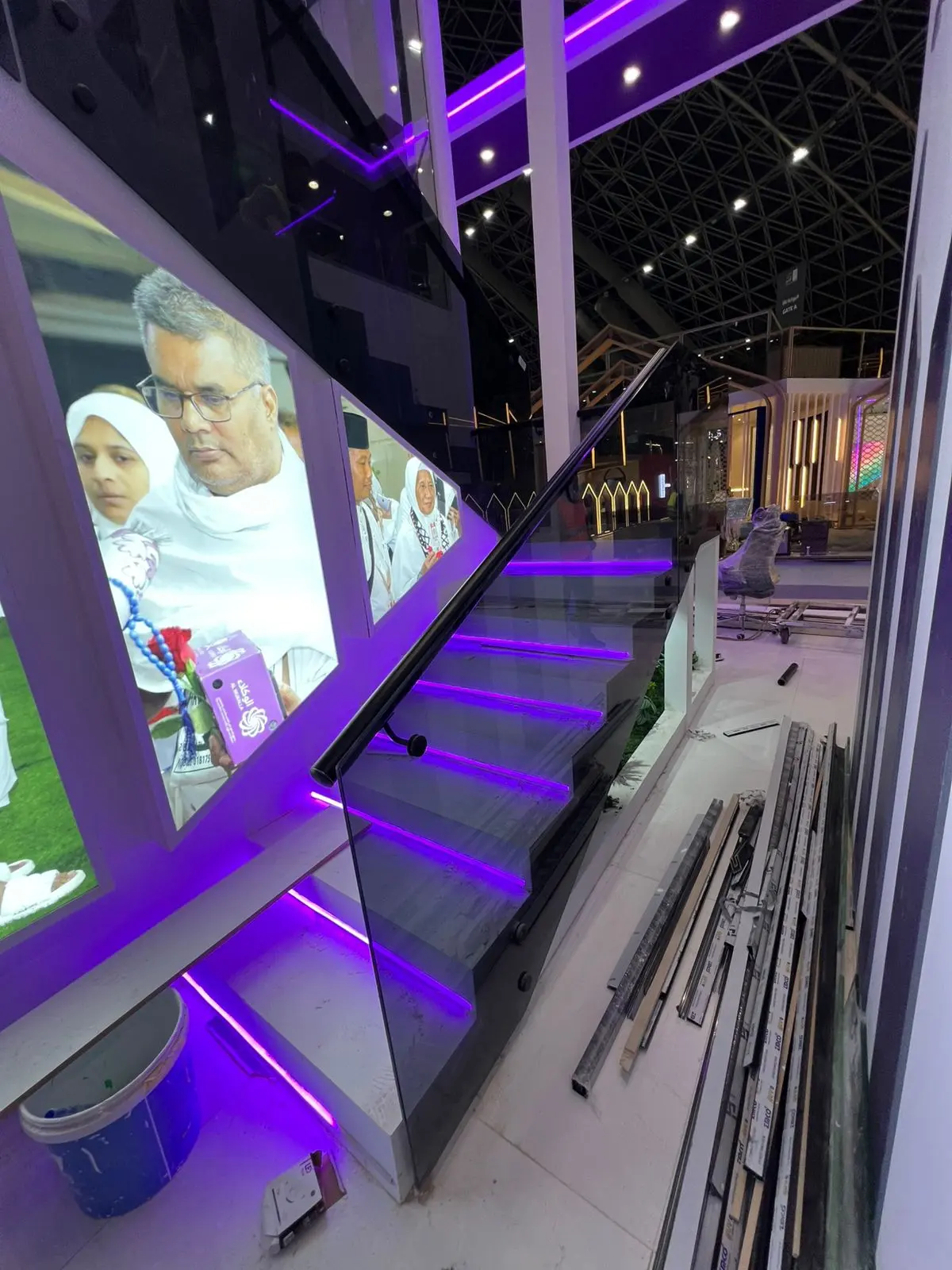 درابزين زجاجي للدرج مثبت بمسامير جانبية (Point Fixing) مع مقبض أسود وإضاءة بنفسجية في جناح معرض لوزارة الحج في جدة.