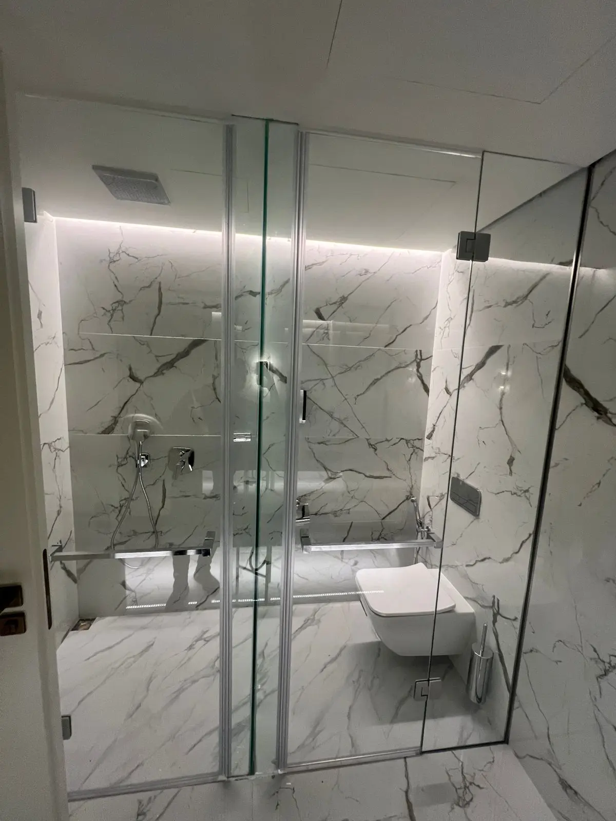 واجهة زجاجية مقسمة لمنطقة الدش والمرحاض بأبواب سيكوريت وإكسسوارات كروم في حمام رخامي مودرن.