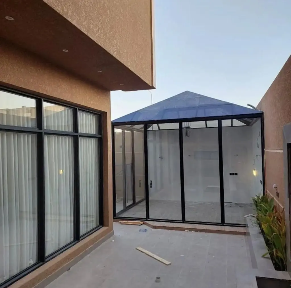 ملحق زجاجي خارجي (Glass House) في فناء منزل بإطارات سوداء وسقف هرمي أزرق.