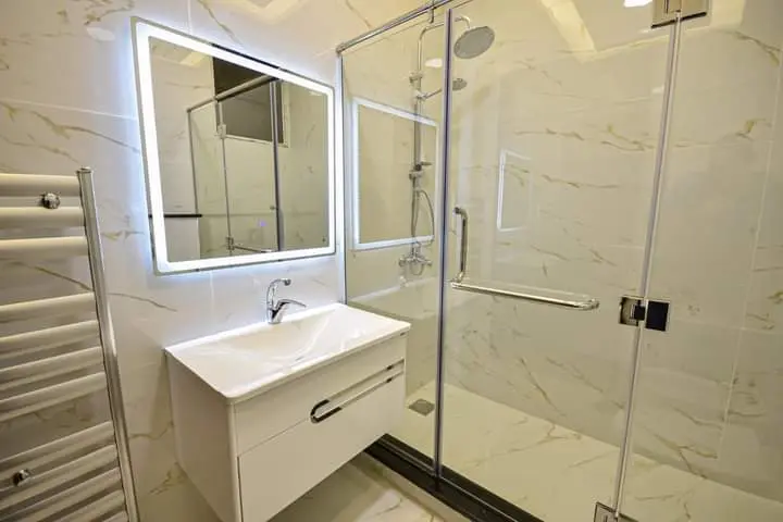 كبينة شاور زجاجية بمفاصل ومقبض كروم بجوار مغسلة مودرن ومرآة بإضاءة خلفية في حمام رخامي.